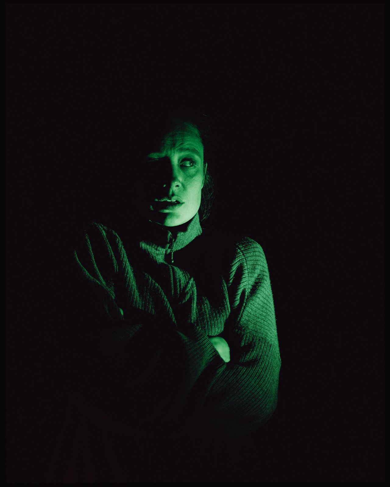 Une femme se tient dans le noir avec une lumière verte et semble avoir très froid.