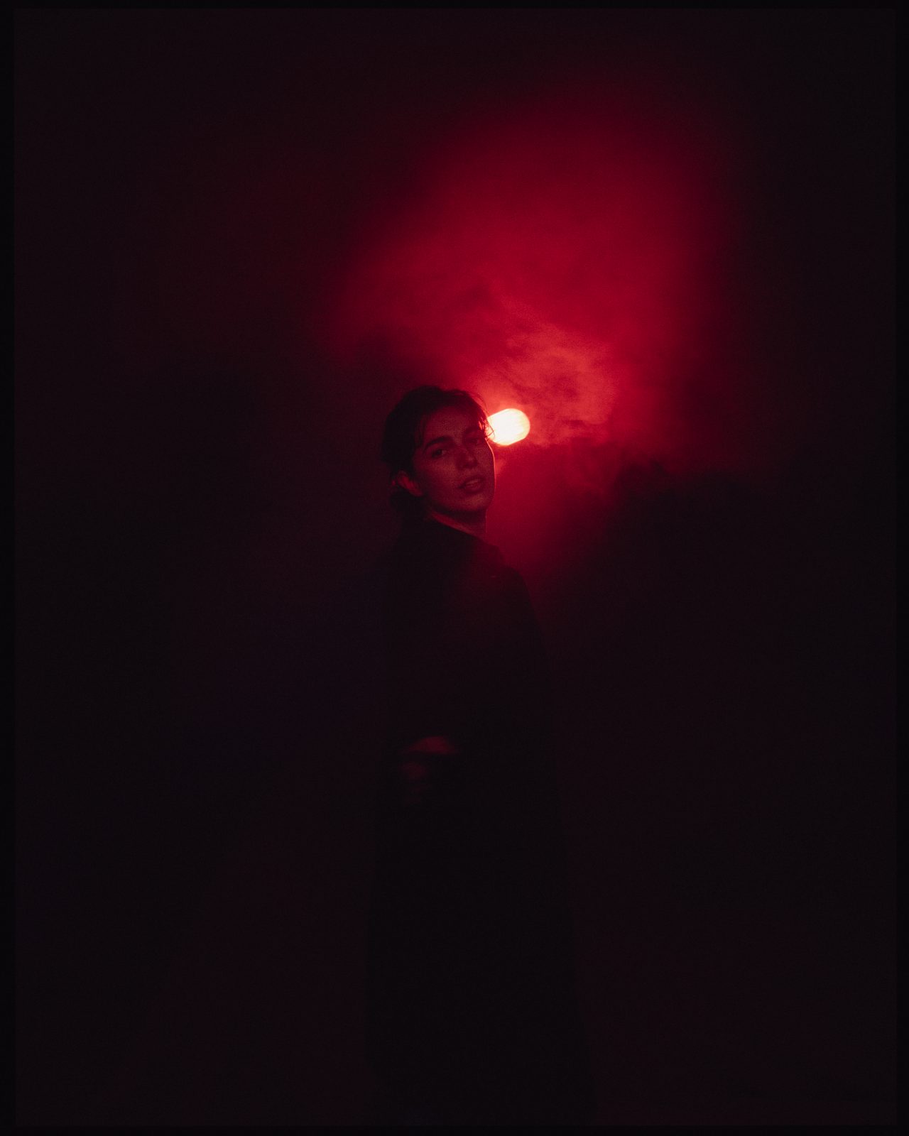 Une femme se tient debout et regarde la caméra, entourée d'une lumière rouge et de fumée.