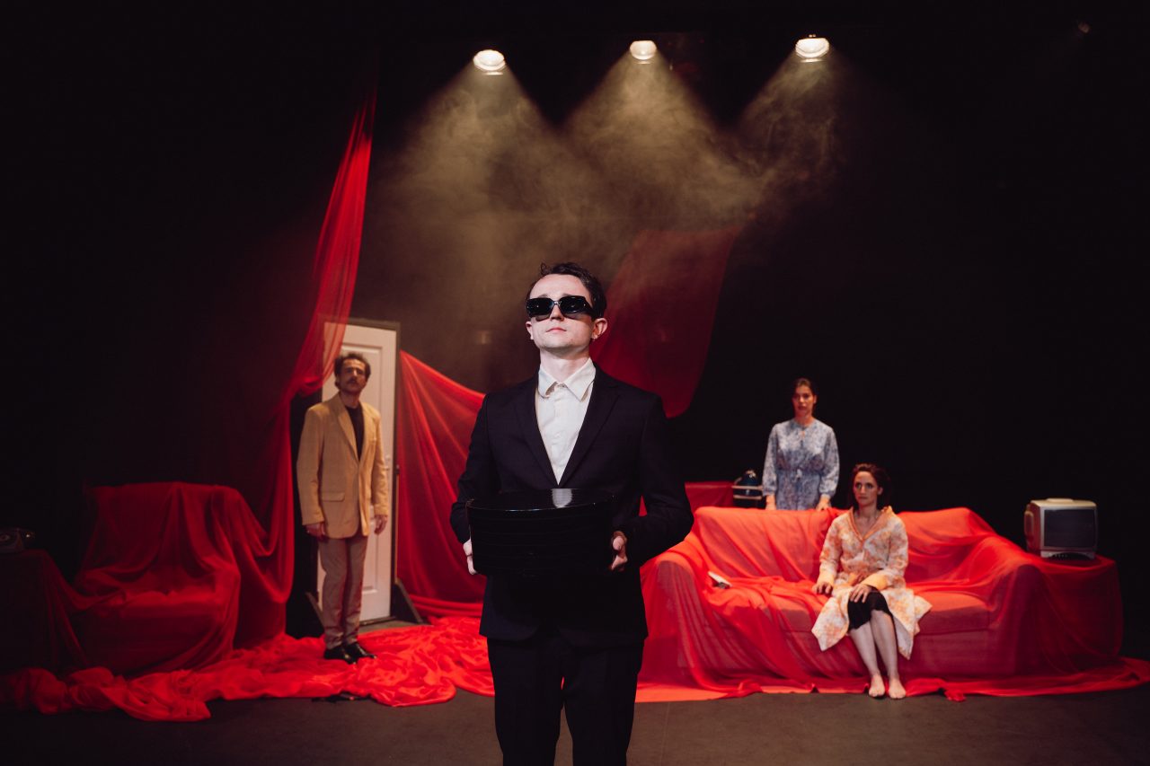 Un homme en costume avec des lunettes noires se tient devant une salle enfumée avec des rideaux rouges. Une femme est assise sur un canapé et une autre femme se tient debout derrière le canapé. Un homme se tient également debout derrière, devant une porte blanche.