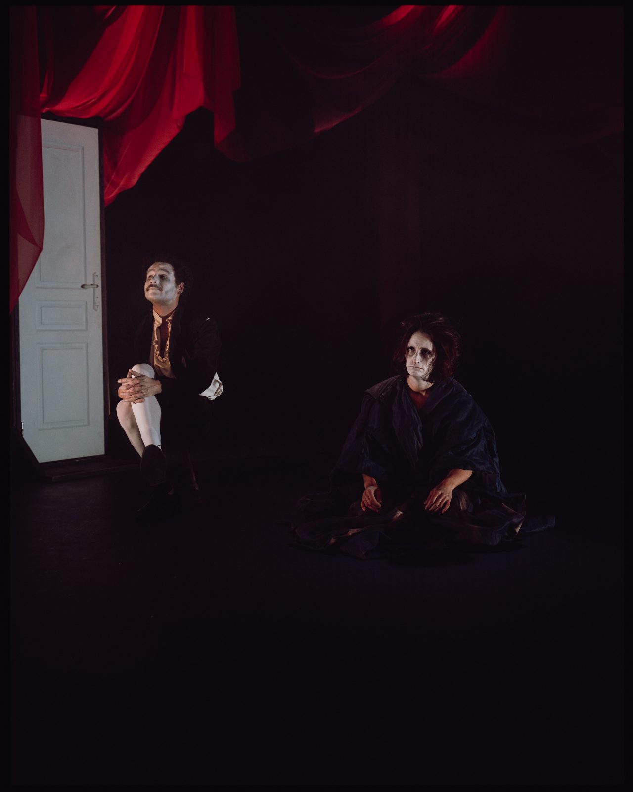 Un homme et une femme maquillé de manière clownesque et effrayante se tiennent dans une pièce, sans se regarder. Une porte blanche se trouve derrière eux.
