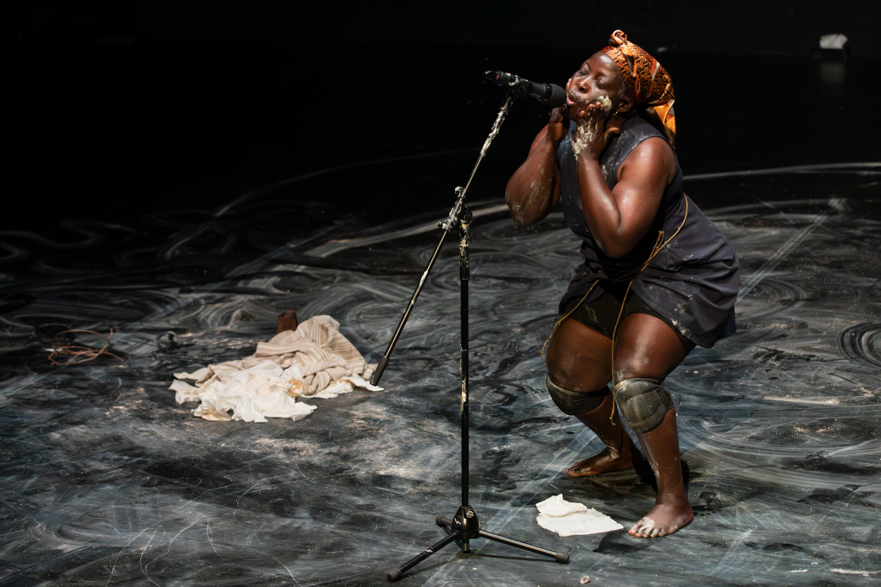 Nadia Beugré chante dans un micro, les genoux pliés, se mettant de la peinture blanche sur le visage.