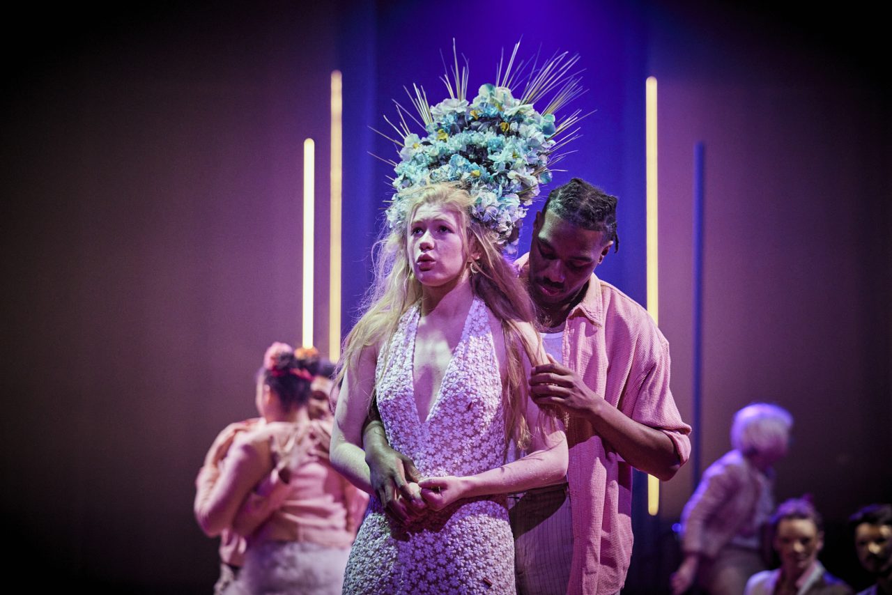 Photos extraite du spectacle. Une femme avec une coiffe de fleurs se tient devant un homme.