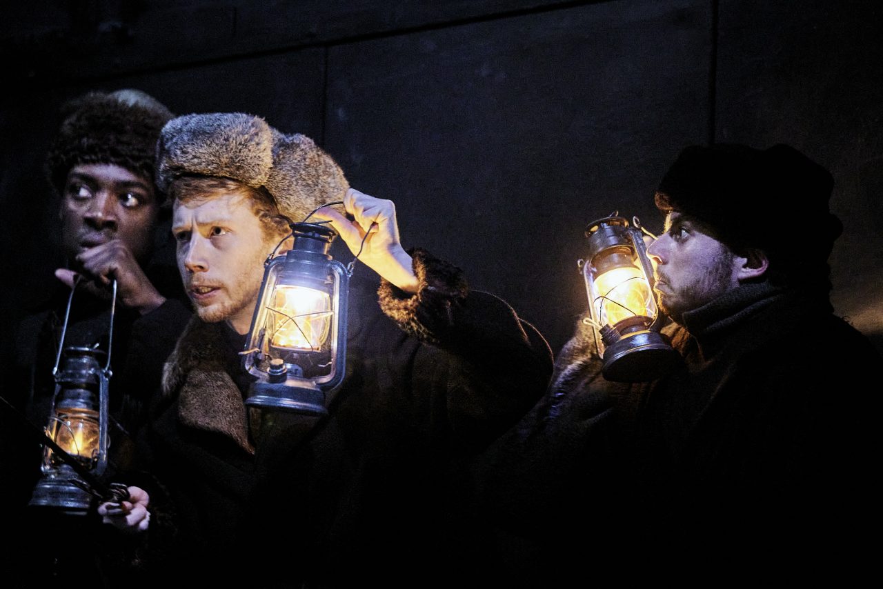 Fond noir. Trois hommes portent des chapeau d'hiver et des lanternes allumées.