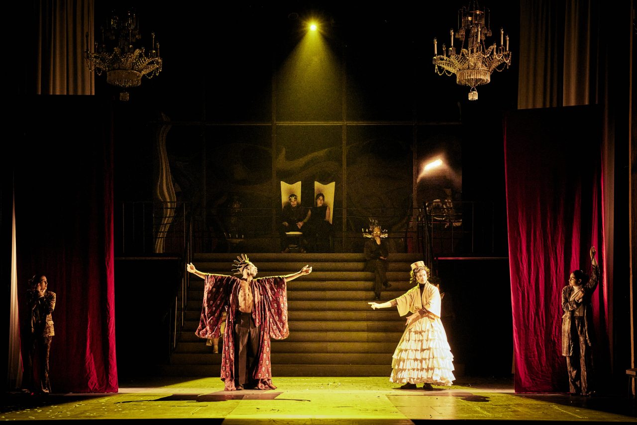 Ambiance lumineuse jeune, deux grand lustres pendent à jardin et à cour. Un personnage habillé en prince se tient devant de grands escaliers, les bras grands ouverts. Un servant se tient à sa gauche. le roi et la reine sont assis sur leur trône en haut des escaliers.